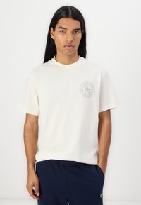 Tricou cu mânecă scurtă, de culoare crem, cu decolteu rotund, având un patch cu logo gri pe pieptul stâng. Material din bumbac neted.