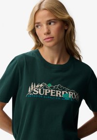 Blondynka w ciemnozielonej koszulce z górami, drzewami, niedźwiedziem i logiem "SUPERDRY Outdoor Supply Co.", spoglądająca w lewo.