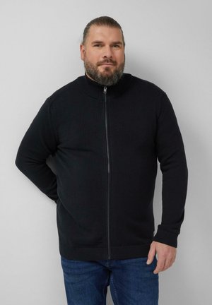 Mężczyzna z brodą i związanymi do tyłu włosami, ubrany w czarny sweter na zamek i niebieskie dżinsy, stoi na tle jednolitego jasnoszarego tła.