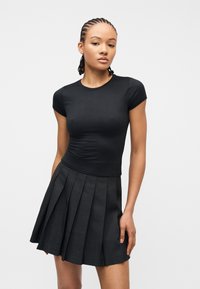 Νέα γυναίκα με πλεγμένα μαλλιά, φορώντας εφαρμοστό μαύρο τοπ με κοντά μανίκια και πλισέ σκουρόχρωμη γκρι φούστα, στέκεται μπροστά σε απλό φόντο.