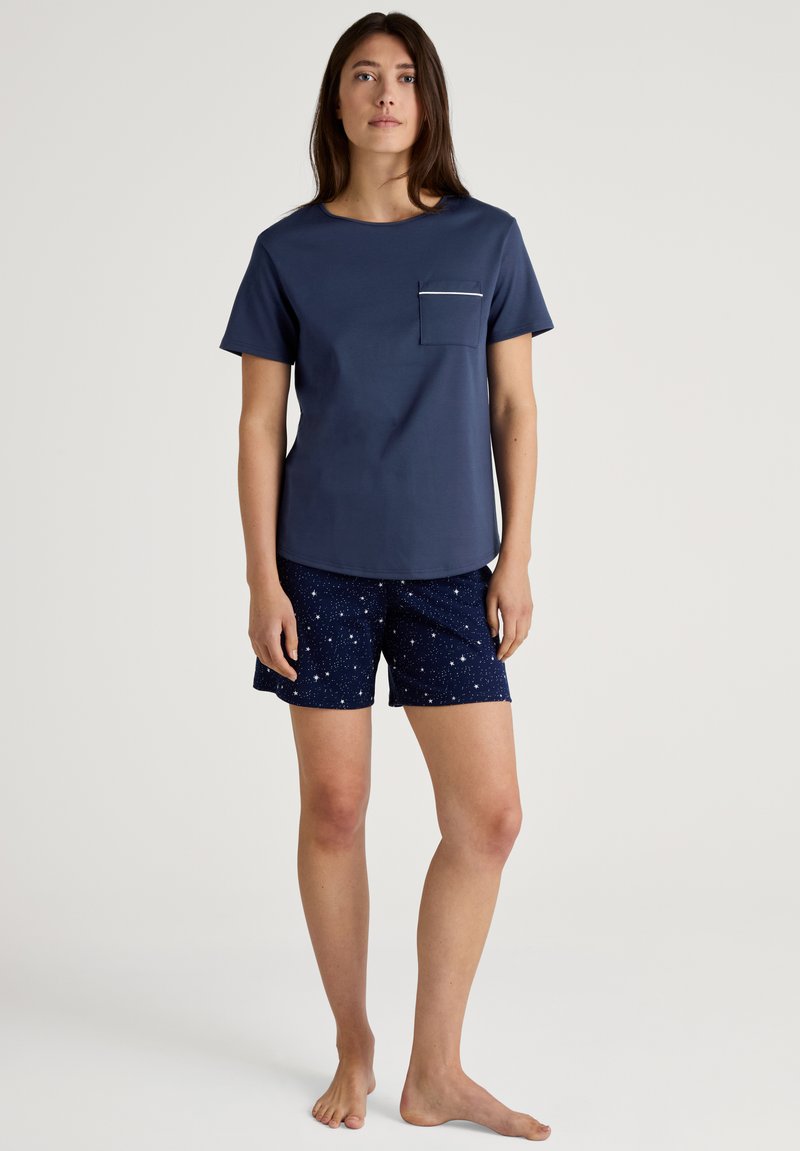 Marineblauwe t-shirt met korte mouwen en een borstzak, gecombineerd met marineblauwe shorts met een sterrenpatroon. Beide zijn gemaakt van zachte stof.