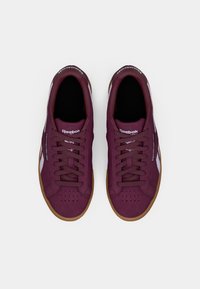 Sapatilhas Reebok em tom bordeaux, com material de camurça, bico redondo, atacadores planos e sola de borracha clara. Apresenta detalhes em branco no logo nas laterais e na língua.