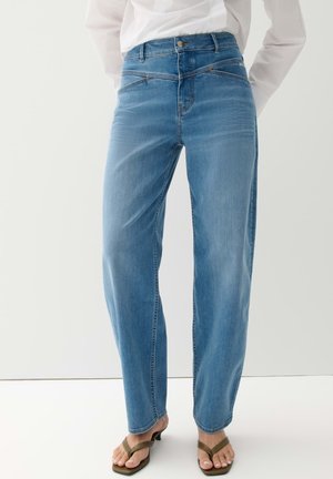 Vrouw die lichtblauwe rechte jeans draagt, een wit shirt met lange mouwen en bruine sandalen met hak, staand tegen een effen achtergrond.
