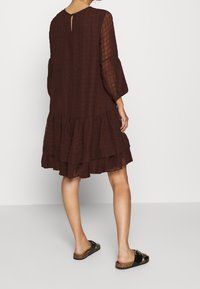 Kvinna klädd i en lös, lager-på-lager mörkbrun klänning med genomskinliga ärmar och svarta slip-on sandaler med korksulor, stående mot en enkel bakgrund.