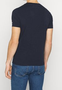 Uomo che indossa una maglietta a maniche corte blu navy e jeans di denim blu, visto da dietro su uno sfondo bianco.