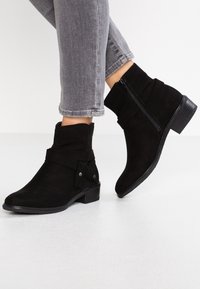 Botas de tobillo de ante negro con un puño doblado, cierre de cremallera lateral y detalle de correa decorativa. Tacón ligeramente elevado y suela de goma.