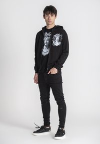 Crna zip-up majica s grafičkim printom, nosi se preko crne majice, uparen s crnim uskim trapericama i crnim tenisicama s bijelim đonom.