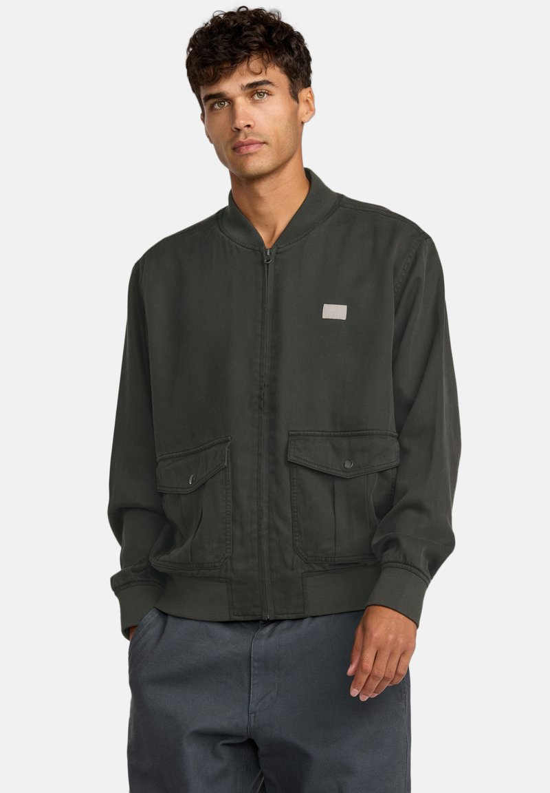 Bomber jacket verde scuro con due tasche frontali, polsini e orlo a coste, e chiusura con cerniera. Presenta un piccolo patch rettangolare con logo.
