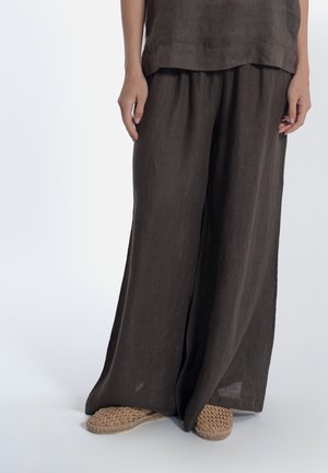 Persona che indossa pantaloni larghi di lino marrone scuro e top abbinato, con scarpe slip-on beige intrecciate, in piedi su sfondo bianco.
