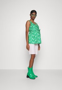 Top floral verde com detalhe em laço, combinado com calções rosa claro. Modelo usa botas de tornozelo verde brilhantes com salto bloco. Fundo minimalista.