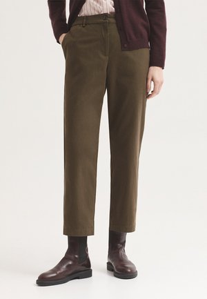 Personne portant un pantalon droit vert olive, des bottines en cuir marron, une chemise rayée et un cardigan violet foncé.