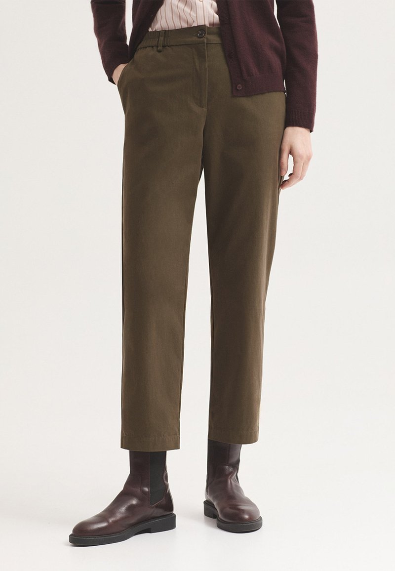 Personne portant un pantalon droit vert olive, des bottines en cuir marron, une chemise rayée et un cardigan violet foncé.