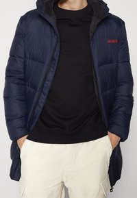 Chaqueta de abrigo azul marino con capucha, que presenta una textura acolchada y el logo rojo "HUGO" en el pecho, superpuesta a un sudadera negra y pantalones beige.
