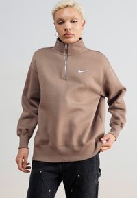 Felpa Nike marrone con colletto alto, zip a un quarto e logo. Presenta maniche lunghe e una vestibilità rilassata, realizzata in tessuto morbido.