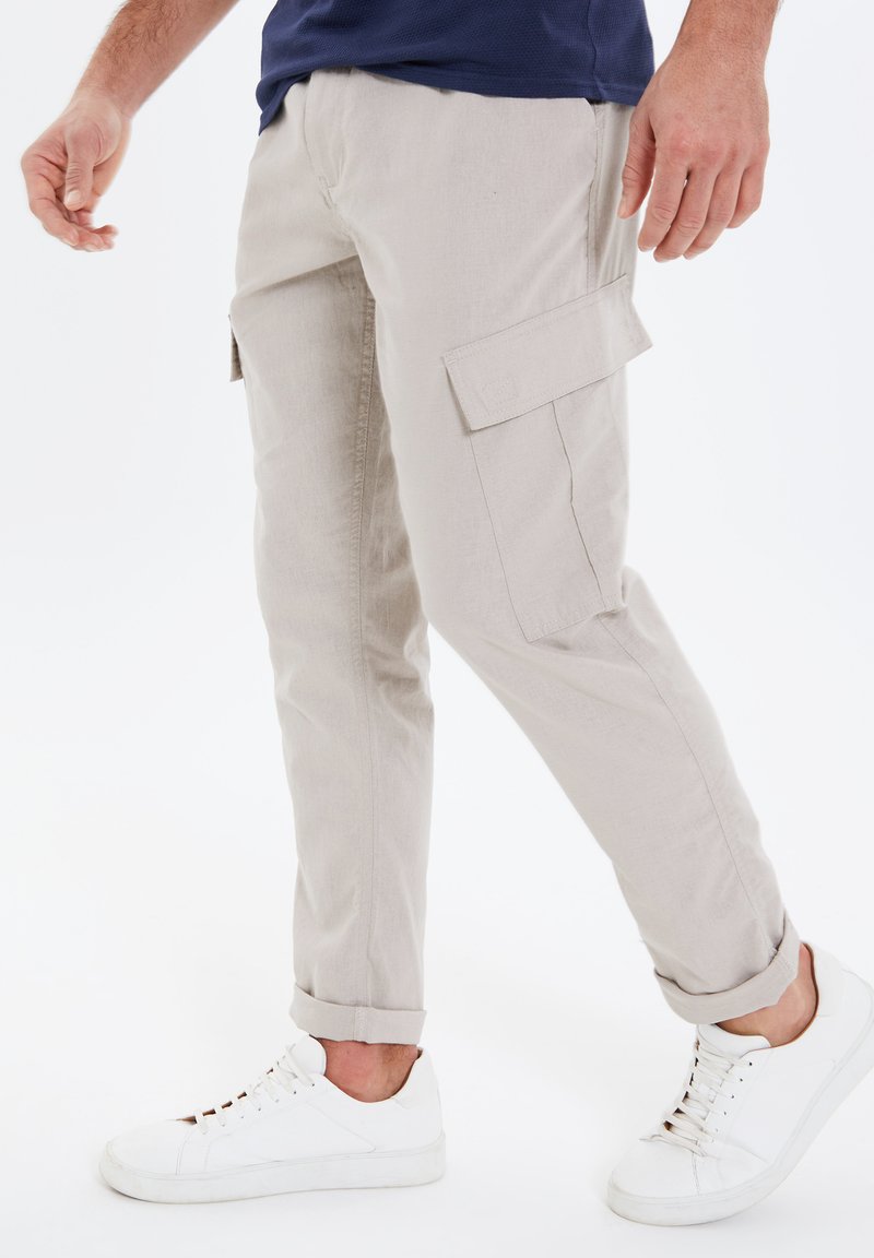 Pantaloni cargo beige con tasche laterali, vestibilità slim, orli arrotolati, realizzati in un tessuto leggero, abbinati a sneaker bianche.