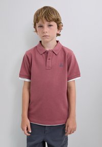 Burgundské polo tričko s texturovanou tkaninou, které má zapínání na dva knoflíky a modrou lebku jako logo. Pod tím jsou bílé krátké rukávy.