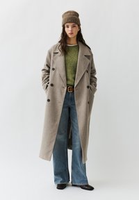 Beige mantel van wolmix met dubbele rij knopen en grote knopen, gedragen over een groene gebreide trui en wijde jeans. Bruine leren riem en gebreide muts.