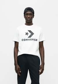 Λευκό βαμβακερό μπλουζάκι με μαύρο λογότυπο Converse και γραφικό αστέρι, συνδυασμένο με μαύρο παντελόνι και μαύρο ριγέ καπέλο.