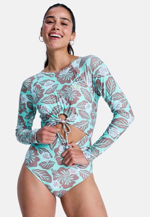 Vrouw glimlachend, draagt een turquoise badpak met lange mouwen en bloemenprint met bruine en witte hibiscussen, past het voorste trekkoord aan.