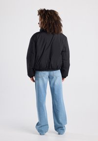 Giacca a vento nera con vestibilità rilassata, vita elasticizzata e polsini a costine. Abbinata a jeans a gamba larga azzurro chiaro, vista da dietro.