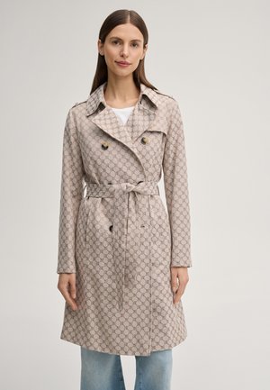 Donna indossa un trench color beige con motivo e cintura annodata in vita sopra jeans azzurri, in piedi contro uno sfondo chiaro e uniforme.