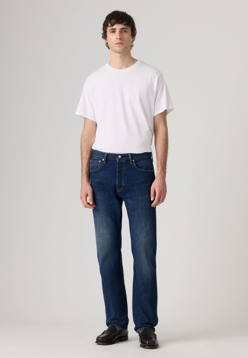 Adult male standing, wearing plain white short-sleeve t-shirt, dark blue jeans, and black dress shoes against a neutral background.