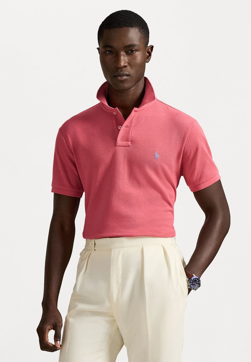 Polo shirt rosa con colletto alto e patta con tre bottoni, abbinato a pantaloni di colore chiaro. Include un piccolo logo blu sul petto.