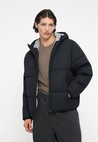 Jachetă puf gri închis cu căptușeală gri la glugă, cu un design matlasat, fermoar frontal și buzunare laterale. Poartă peste o bluză maro și pantaloni gri.