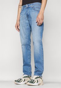 Jeans din denim de culoare albastru deschis, cu croi drept, având un design cu cinci buzunare și decolorare subtilă, asortați cu pantofi sport verzi și crem.