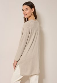 Beige långärmad tunika med sidoklyftor, mjuk textur och rund halsringning, tillsammans med ljusa byxor. Minimalistisk design.