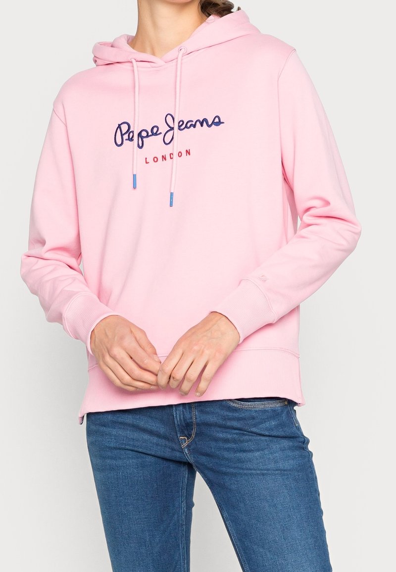 Personne portant un sweat à capuche rose avec le texte "Pepe Jeans London", associé à un jean bleu, les mains jointes devant.