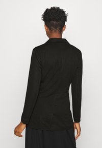 Blazer noir à manches longues, col cranté, en tissu texturé. Caractéristiques : coupe ajustée et fente arrière pour faciliter les mouvements.