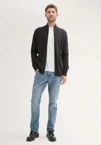 Sötét, cipzáras pulóver fehér póló fölött, világoskék farmernadrággal és fekete cipővel párosítva. Simább anyagból, testhezálló szabással.