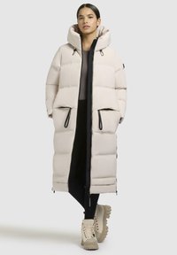 Long, light beige puffer coat with a hood, dual zip pockets, and black zipper accents. Smooth texture and oversized fit.