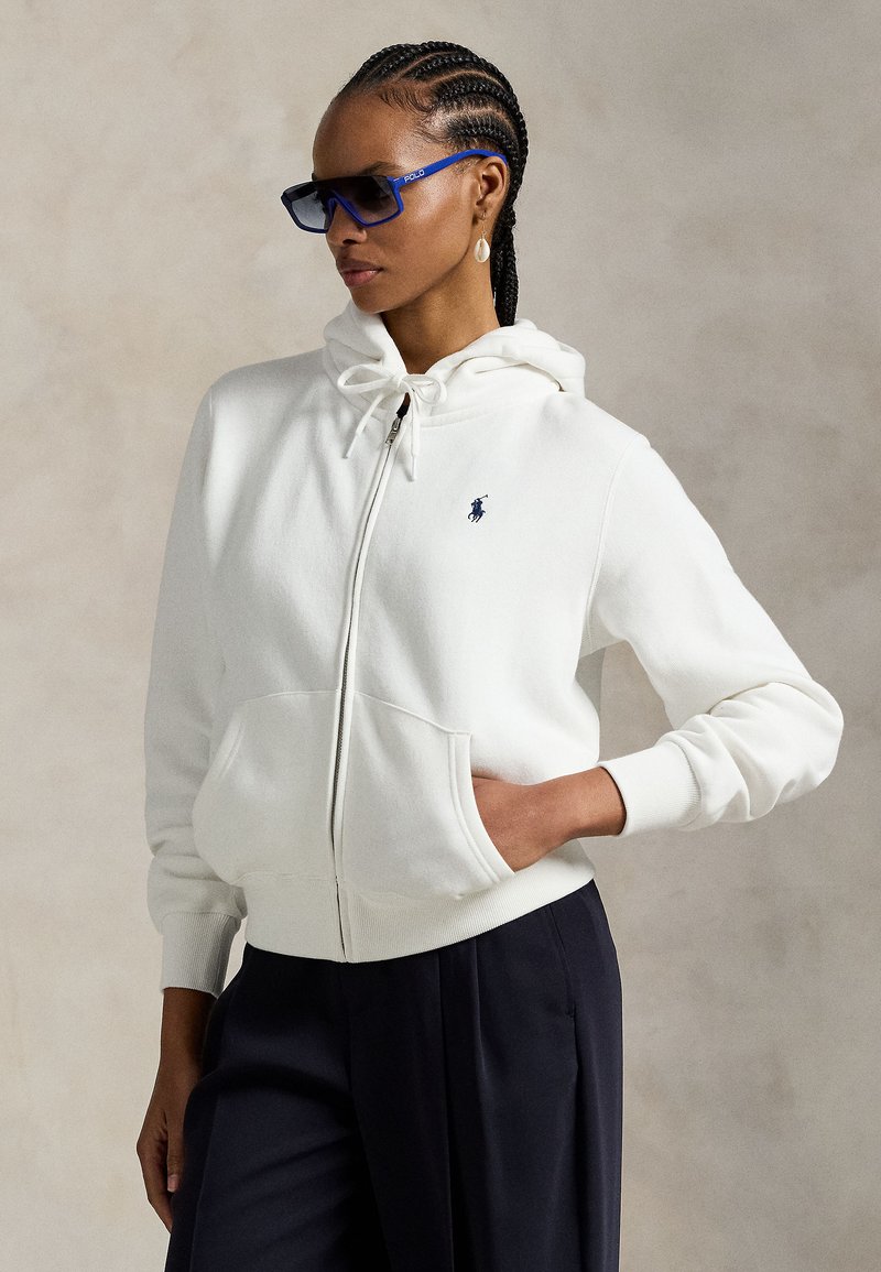 Femme aux cheveux tressés portant des lunettes de soleil bleues, un sweat à capuche blanc zippé avec poche et logo, et un pantalon foncé, posant devant un fond neutre.