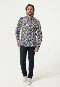 Overhemd met knopen en een meerkleurig bladerenpatroon in blauw, groen en bruin op een witte achtergrond. Donkere spijkerbroek en witte sneakers.