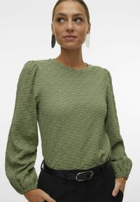Camicetta verde a maniche lunghe con spalle a sbuffo, scollatura a costine e cintura nera. Il tessuto presenta un motivo tessuto a pois.