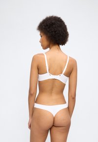 Γυναίκα με κατσαρά μαλλιά που φοράει λευκό σουτιέν και ταίριαστο στρινγκ, εμφανίζεται από πίσω σε απλό φωτεινό φόντο.