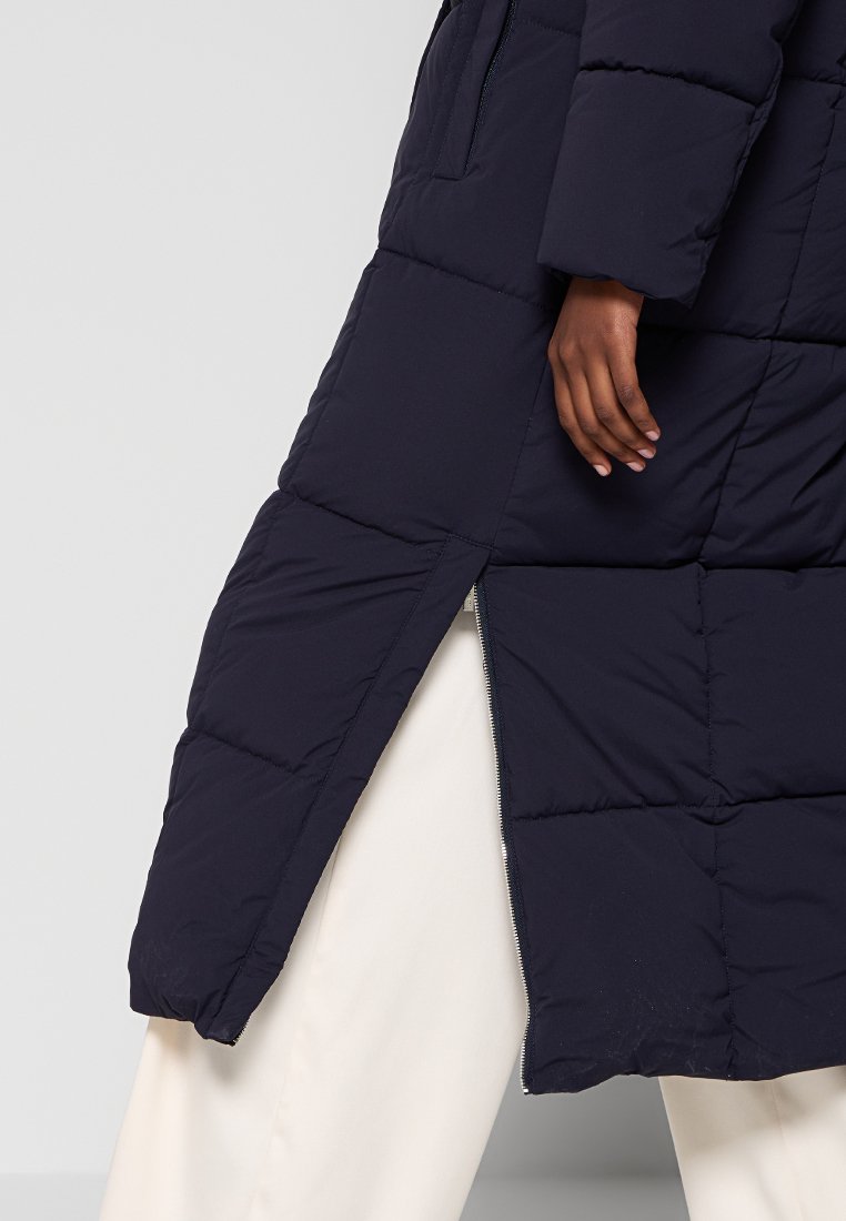 Cappotto lungo puffer blu navy, design trapuntato con spacco frontale, chiusura con zip e tasche laterali. Il tessuto è liscio e ha una consistenza morbida.