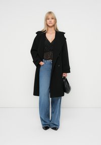Blondynka w czarnym, oversize'owym płaszczu, przezroczystej bluzce w czarne kropki, niebieskich szerokich dżinsach, czarnych szpiczastych butach, trzymająca czarną torebkę.