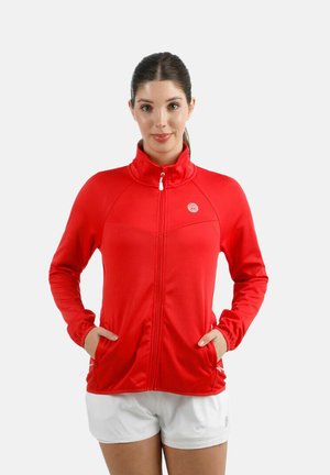 Vrouw met bruin haar in een staart, gekleed in een rode jas met rits en een witte korte broek, met haar handen in de jaszakken, staand tegen een witte achtergrond.