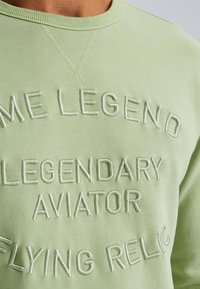 Svijetlozeleni dukserica s reljefnom vezom s natpisom "NEKI LEGENDA LEGENDARNI AVIATOR FLYING RELIC", okrugli izrez, i mekana tekstura.