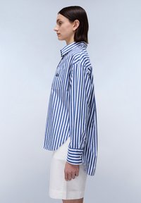 Camicia a righe in cotone blu e bianco con colletto button-down, taschino sul petto e spacco laterale; abbinata a pantaloni corti bianchi.