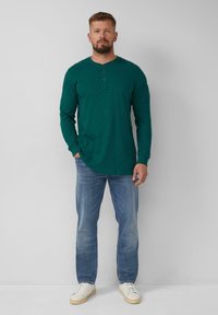 Teal Langarm-Henley-Shirt mit drei Knöpfen, kombiniert mit hellblauen Jeans und weißen Sneakers. Das Shirt hat eine strukturierte Oberfläche und einen figurbetonten Schnitt.