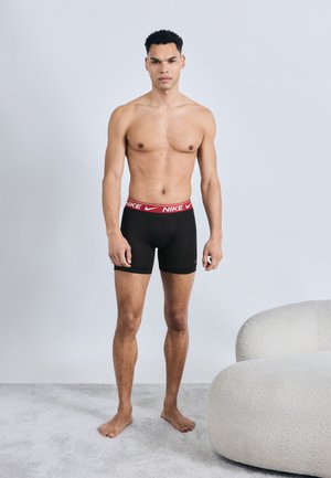 Άνδρας που στέκεται ξυπόλητος σε γκρι χαλί φορώντας μαύρα boxer briefs της Nike με κόκκινη μέση, δίπλα σε λευκή στρογγυλεμένη πολυθρόνα μπροστά σε γκρι απλό τοίχο.