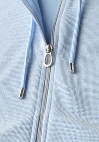 Felpa con zip di colore blu chiaro, realizzata in un tessuto strutturato, con zip argentata e coulisse abbinate con punte metalliche. Vista ravvicinata della zona della zip.