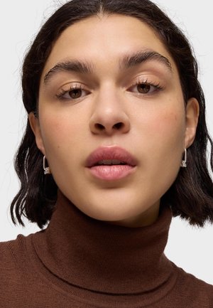 Joven mujer con cabello corto y oscuro, lleva pendientes plateados de forma cuadrada y un jersey de cuello alto marrón, expresión neutral, mirando al frente.