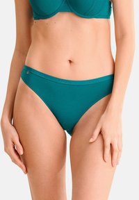 Femme portant un soutien-gorge turquoise et une culotte de style bikini assortie, debout avec les mains reposant sur les cuisses devant un fond uni.