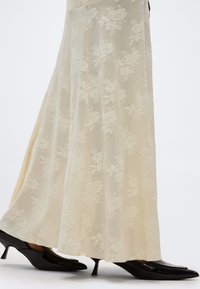 Μακρύ κρεμ δαντελένιο φόρεμα με λουλουδάτο μοτίβο, συνδυασμένο με μαύρα μυτερά παπούτσια με μικρό τακούνι, εμφανιζόμενο από το μέσο της γάμπας και κάτω.