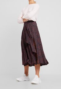 Schwarzer Rock mit roten Pünktchen, asymmetrischem Rüschensaum und seitlichem Schlitz, kombiniert mit weißen Sneakern und einem hellrosa Langarmoberteil.